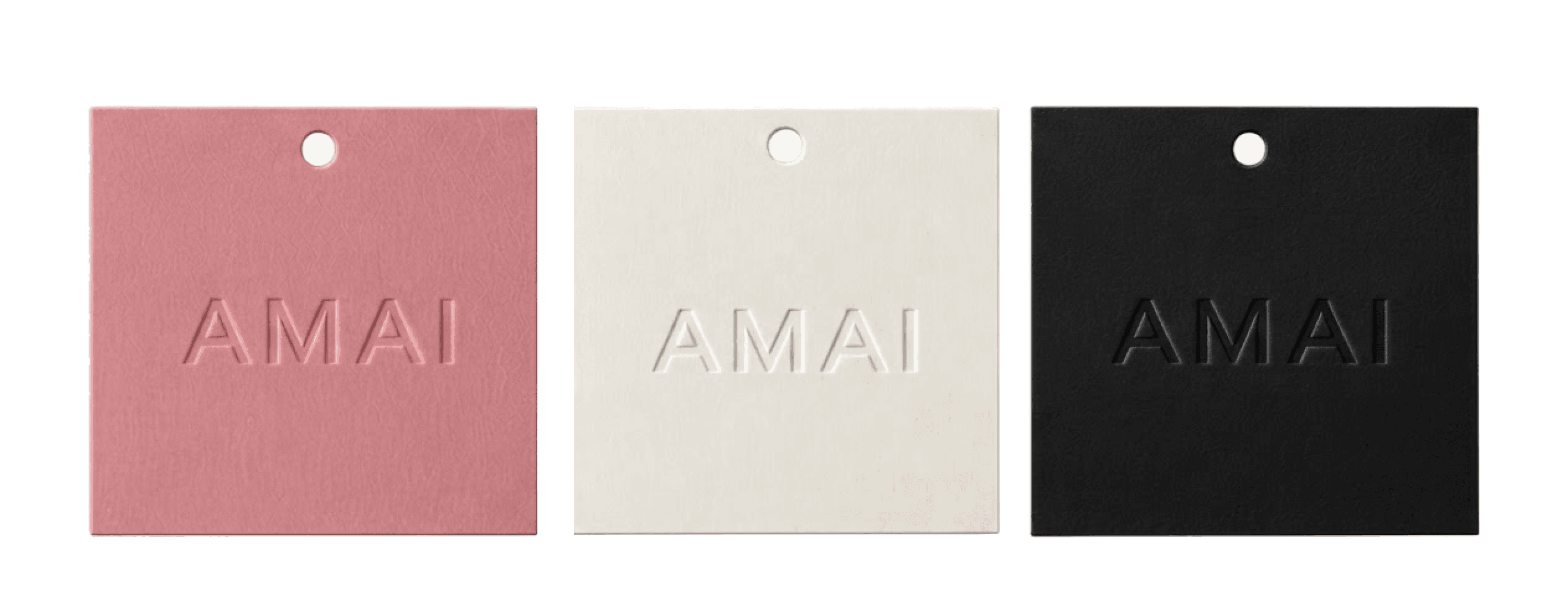 Three square AMAI hang tags in dusty pink, cream and black, each with an embossed wordmark and a punched hole.